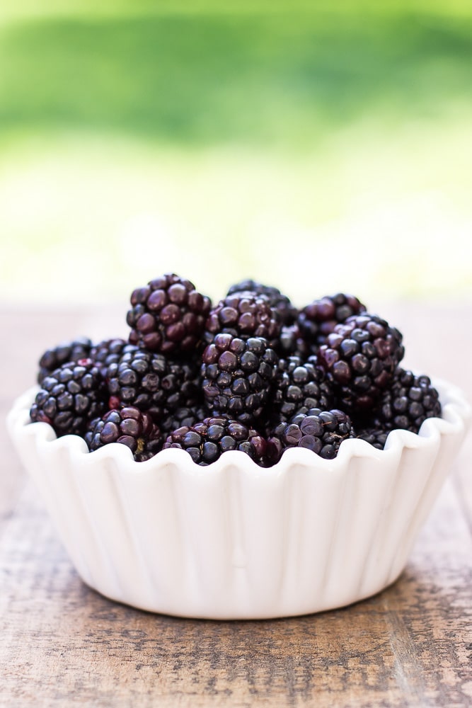 A white bowl of blackberries on a wooden board