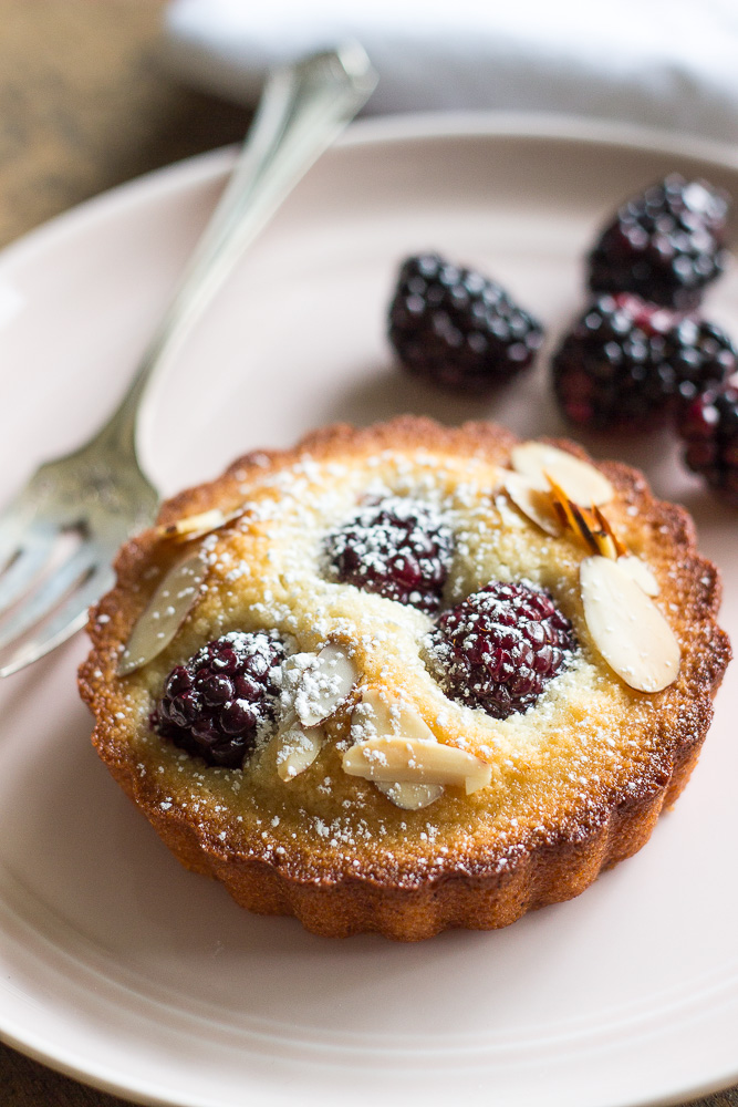 An almond blackberry financier on a pink plate with a fork and a few blackberries