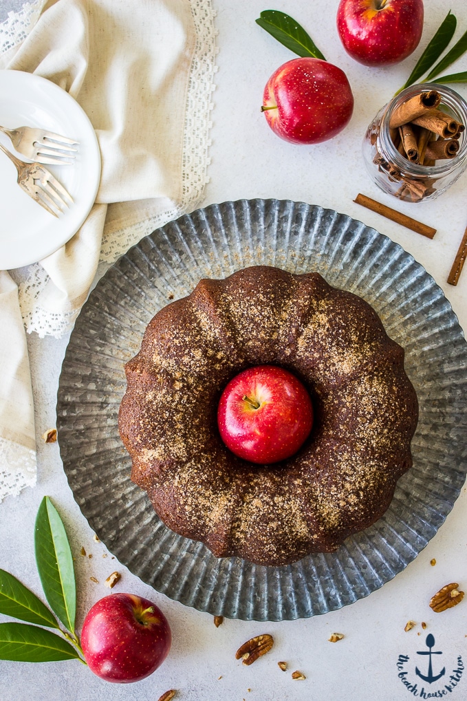 Overhead photo of Apple Cider Doughnut Bundt Cake with Pecan Brown Sugar Ripple with a red apple in middle of cake on a silver tray surrounded by red apples and cinnamon sticks.