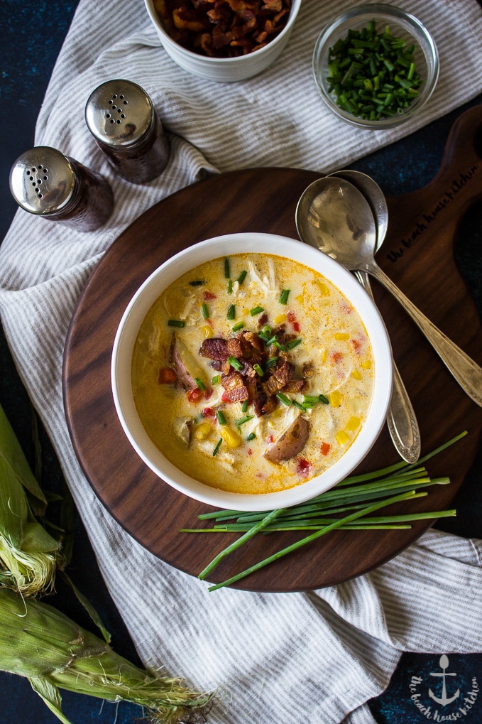 Overhead photo of Creamy Chicken Corn Chowder with Bacon in a white bowl, topped with bacon and chives on a dark brown board with green chives in front.