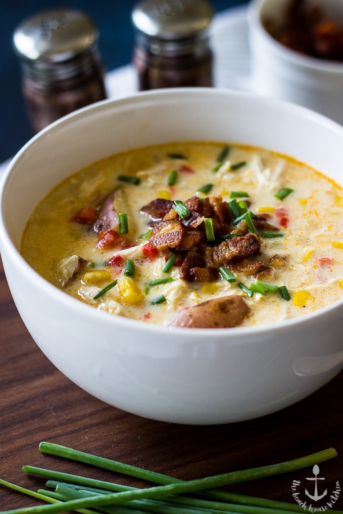 Creamy Chicken Corn Chowder with Bacon in a white bowl, topped with bacon and chives on a dark brown board with green chives in front.