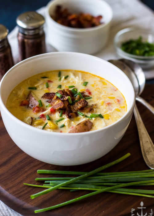Creamy Chicken Corn Chowder with Bacon in a white bowl with bacon and chives on top, on a dark wood tray with chives in front of bowl.