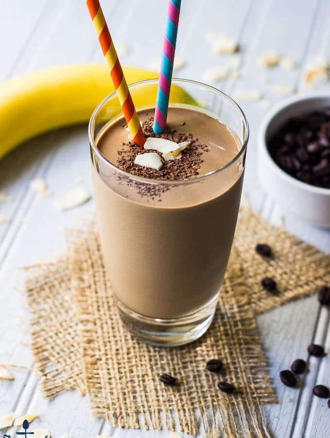Chocolate Coconut Coffee Smoothie with two colorful straws, on top of two pieces of burlap ribbon with a banana in background.
