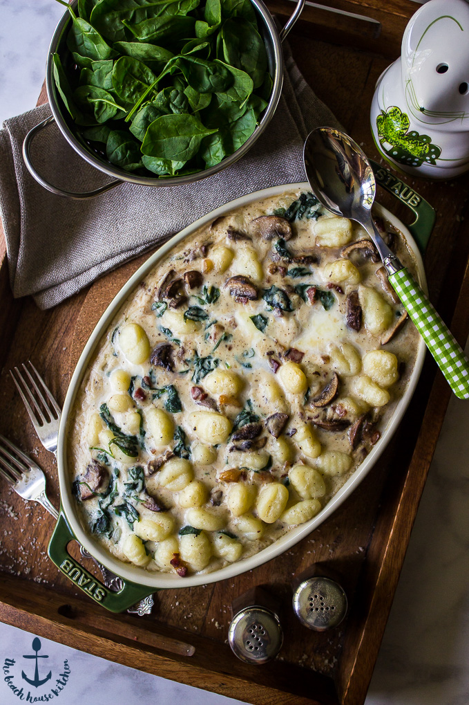 An oval dish filled with Mushroom Florentine Gnocchi on a wooden tray with a colander of fresh spinach.