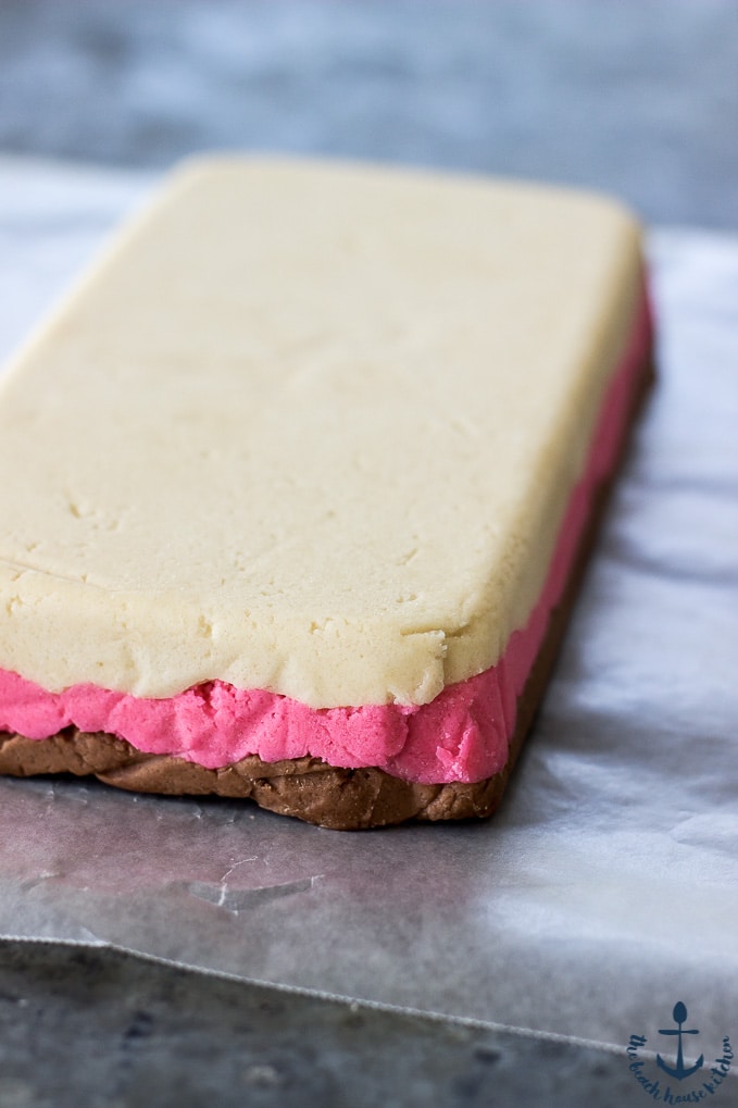 A slab of Neapolitan Butter Cookie dough stacked white, pink, chocolate on a piece of wax paper.