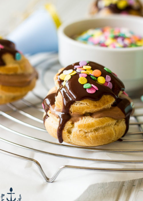 Nutella Cream Puffs drizzled with chocolate and sprinkles on a round silver cooling rack with a white bowl of sprinkles in in center of rack.