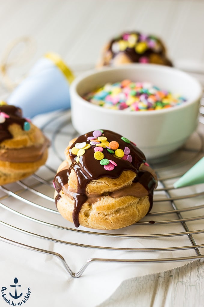 Nutella Cream Puffs drizzled with chocolate and sprinkles on a round silver cooling rack with a white bowl of sprinkles in in center of rack.