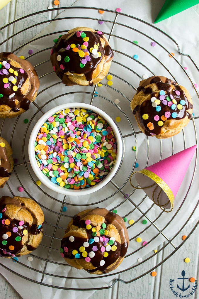 Overhead photo of Nutella Cream Puffs drizzled with chocolate and sprinkles on circular silver cooling rack with white bowl of sprinkles in center with mini party hats.