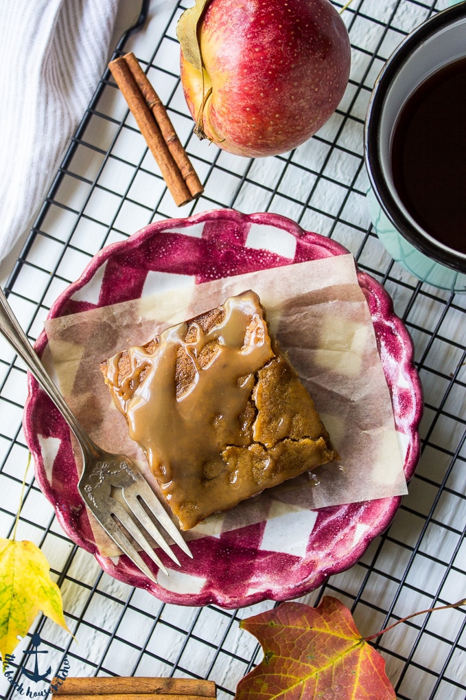 A Caramel Glazed Apple Butter Blondie on a red and white check plate with a fork, an apple, a few fall leaves on top of a cooling rack.