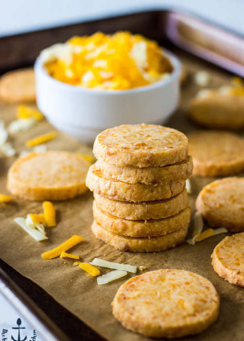 Stack of Cheddar Cheese Coins on baking sheet lined with parchment with white bowl filled with shredded cheddar cheese.