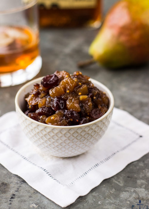 Pear Cranberry Bourbon Chutney in a small white bowl, on a white napkin with a pear and glass of bourbon in background.