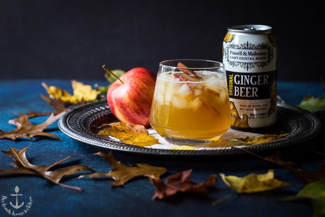 The Fall Dark 'N' Stormy cocktail on a silver tray with an apple and a can of ginger beer with leaves surrounding tray.