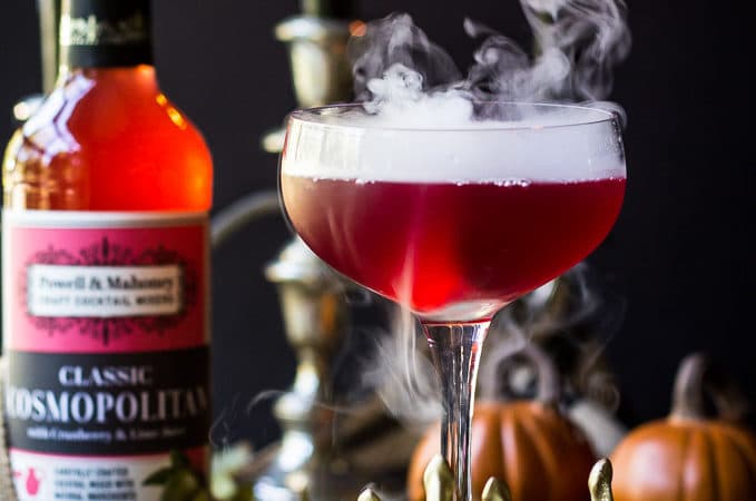 Red Vampire's Venom Cocktail with dry ice smoke bubbling in a gold skeleton hand with candles and cocktail bottle in background.