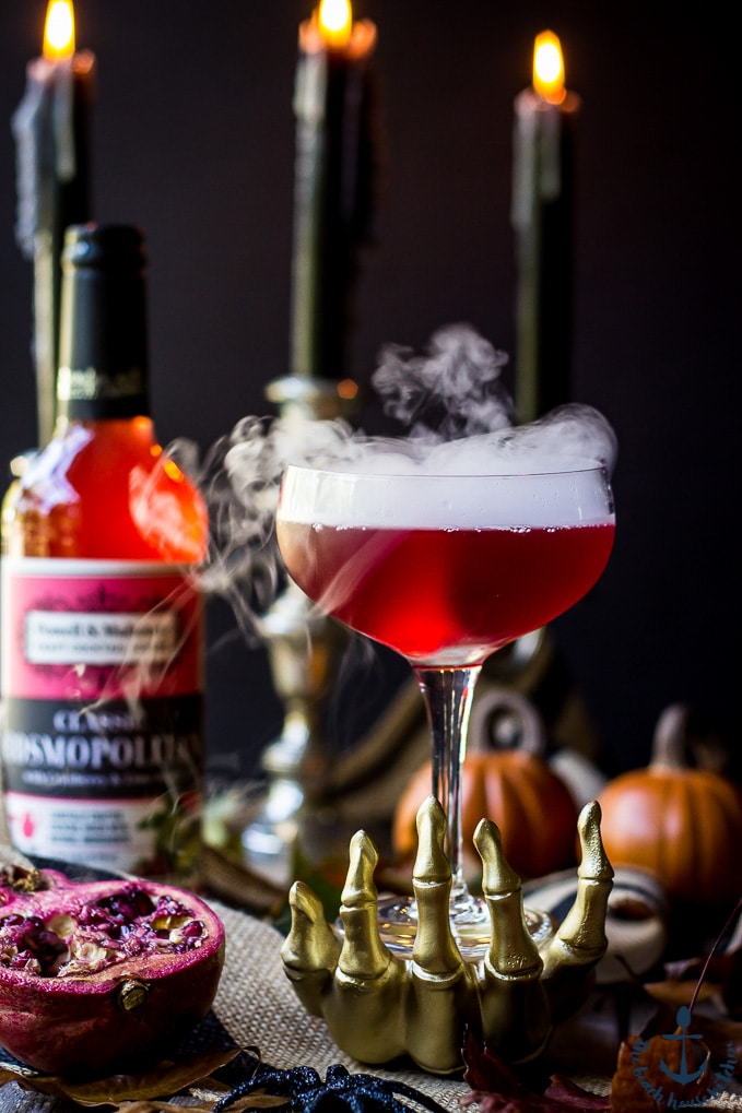 Red Vampire's Venom Cocktail with dry ice smoke bubbling in a gold skeleton hand with candles and cocktail bottle in background.