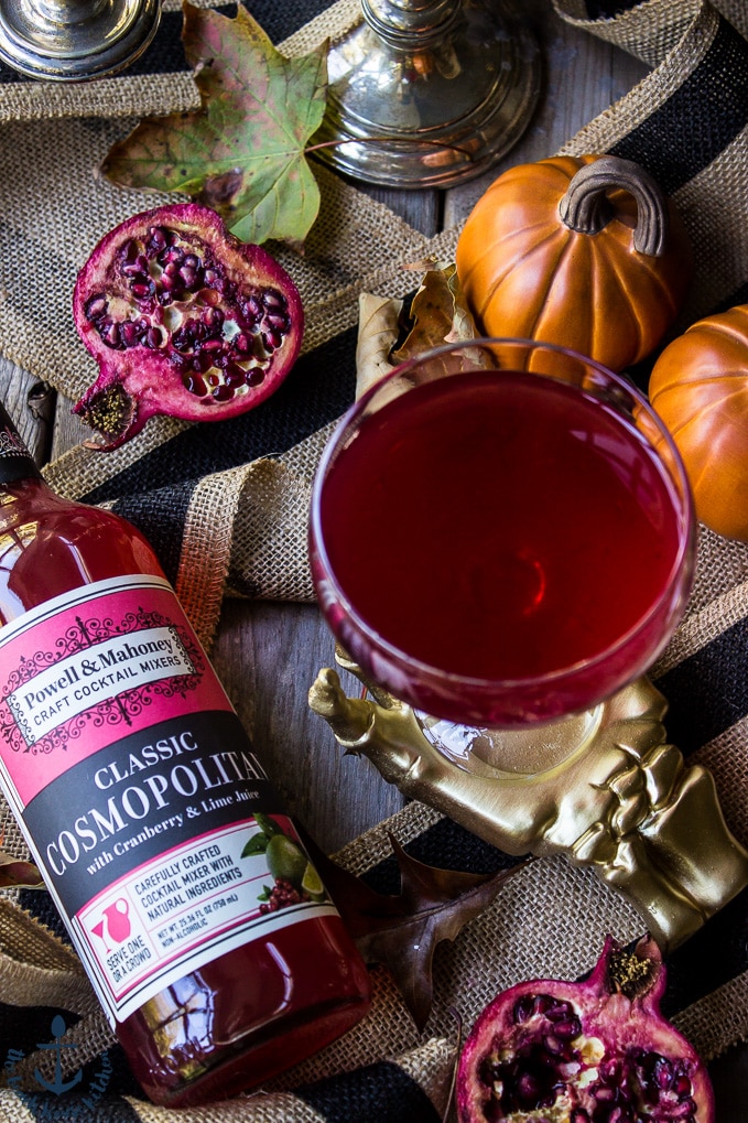 Overhead photo of glass filled with red cocktail and a bottle of cosmopolitan mixer, mini pumpkins and a half a pomegranate.
