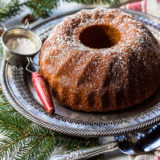 Honey Pecan Bundt Cake on silver tray with greens, cake knife, sifter, honey and lemon in background.