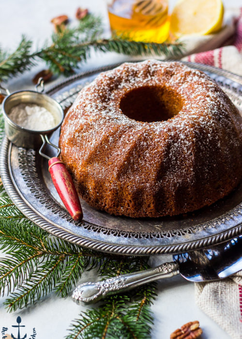 Honey Pecan Bundt Cake on silver tray with greens, cake knife, sifter, honey and lemon in background.