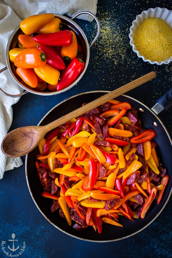 Red and yellow pepper strips in frying pan along with colander of mini pepper and white bowl of dried polenta.