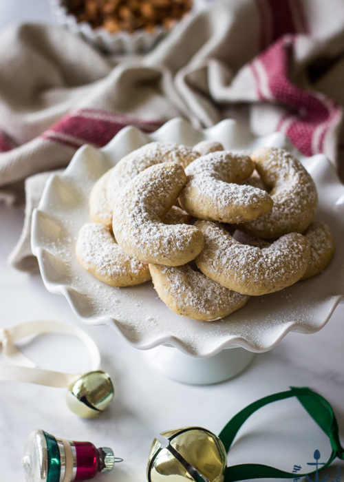 Almond Crescents dusted with confectioners' sugar on white pedestal plate with jingle bells in background.
