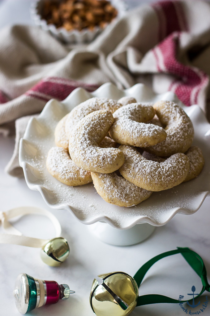 Almond Crescents dusted with confectioners' sugar on white pedestal plate with jingle bells in background.