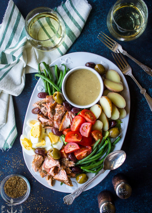 Overhead photo of Salmon Nicoise Salad with Dijon Vinaigrette on a blue board surrounded by wine glasses, salt and pepper shakers, forks and spices.
