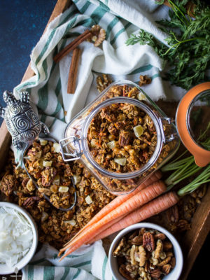 Overhead shot of carrot cake granola in jar on try with bowls of nuts and coconut, carrots, and scooper filled with granola.