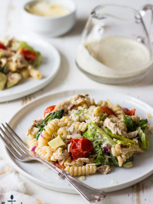 A round, white plate topped with a pasta salad filled with tomatoes, greens and spiral noodles with a silver fork on a white wood surface.
