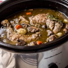 Coq Au Vin in a silver slow cooker with a bottle of wine and salt and pepper shakers in background.