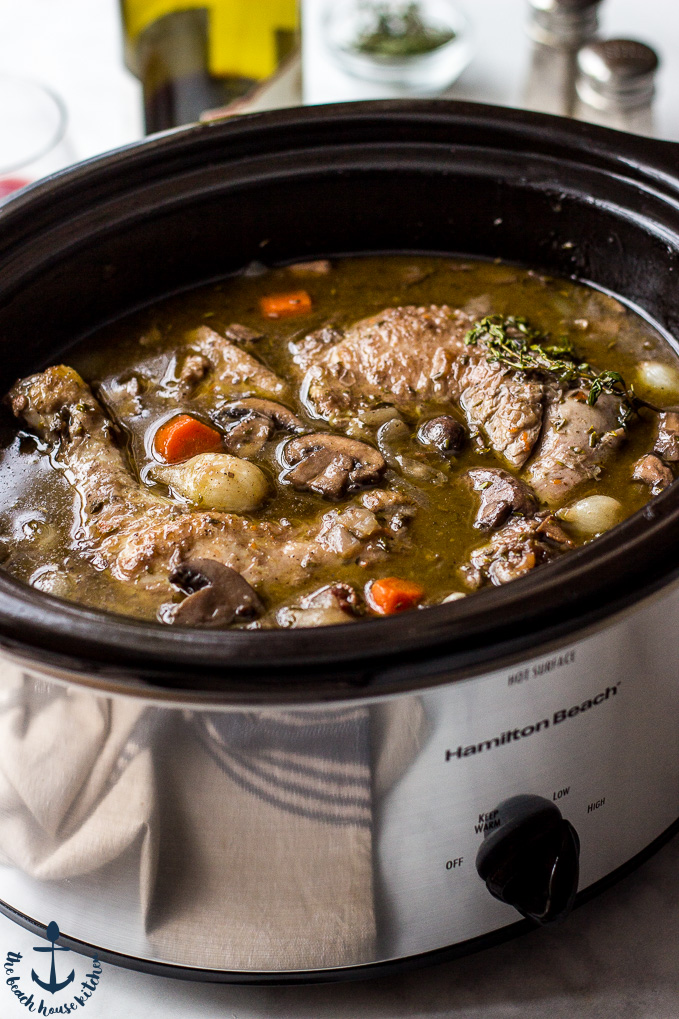 Slow Cooker Coq Au Vin in a silver slow cooker with a bottle of wine and salt and pepper shakers in background.