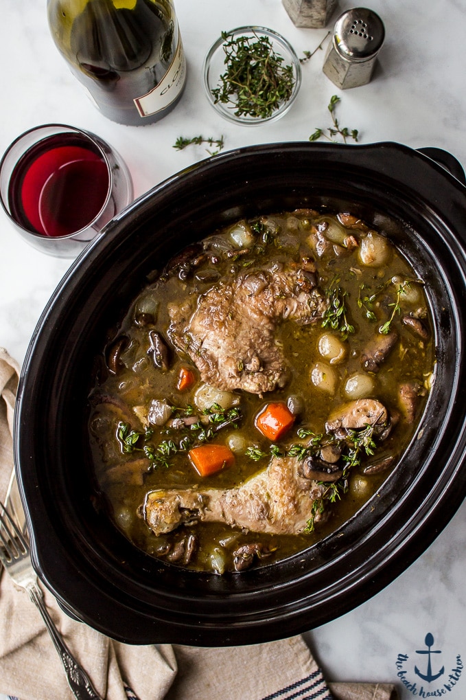 Overhead photo of Slow Cooker Coq Au Vin in a black slow cooker with a bottle of wine , glass of wine and salt and pepper shakers in background.
