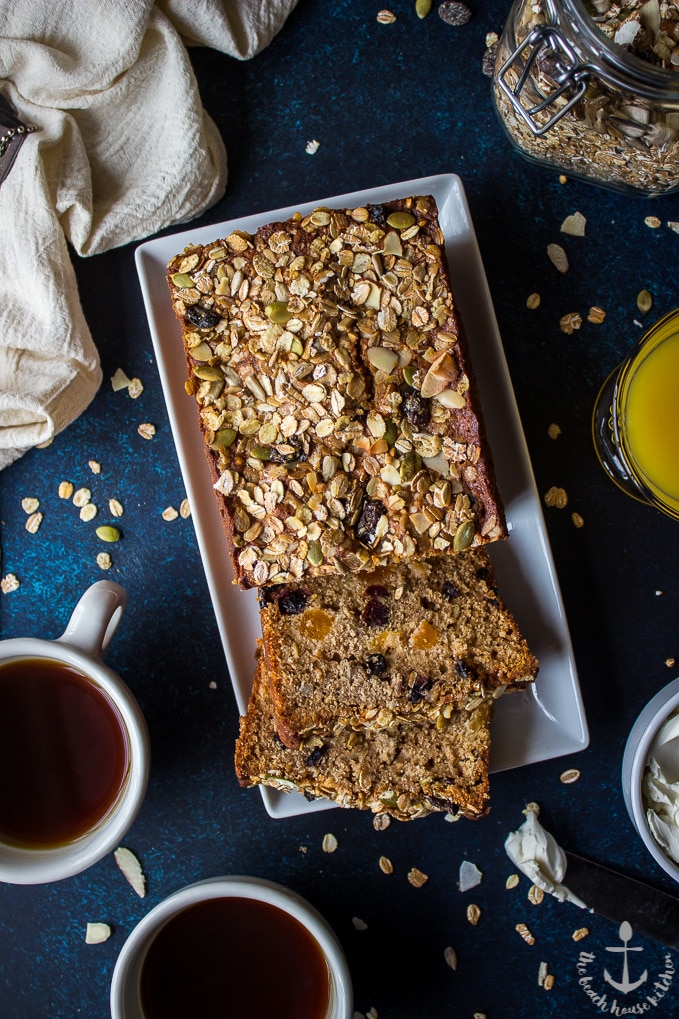 Overhead shot loaded muesli quick bread on blue board with two coffee cups, glass of orange juice and jar of muesli.