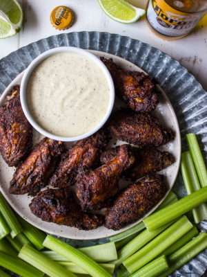 Overhead shot of spicy baked spicy wings with white bbq sauce on tray with celery and beer and lime in background.