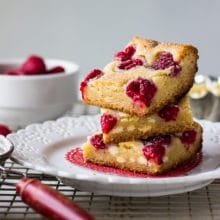 Stack of white chocolate raspberry bars on white plate on top of wire rack with raspberries in bowl in background.