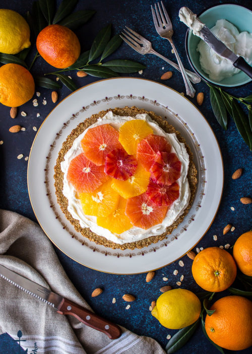 Overhead shot of winter citrus mascarpone tart on white plate crust with oranges, lemon, sharp knife, forks and bowl filled with whipped cream in background