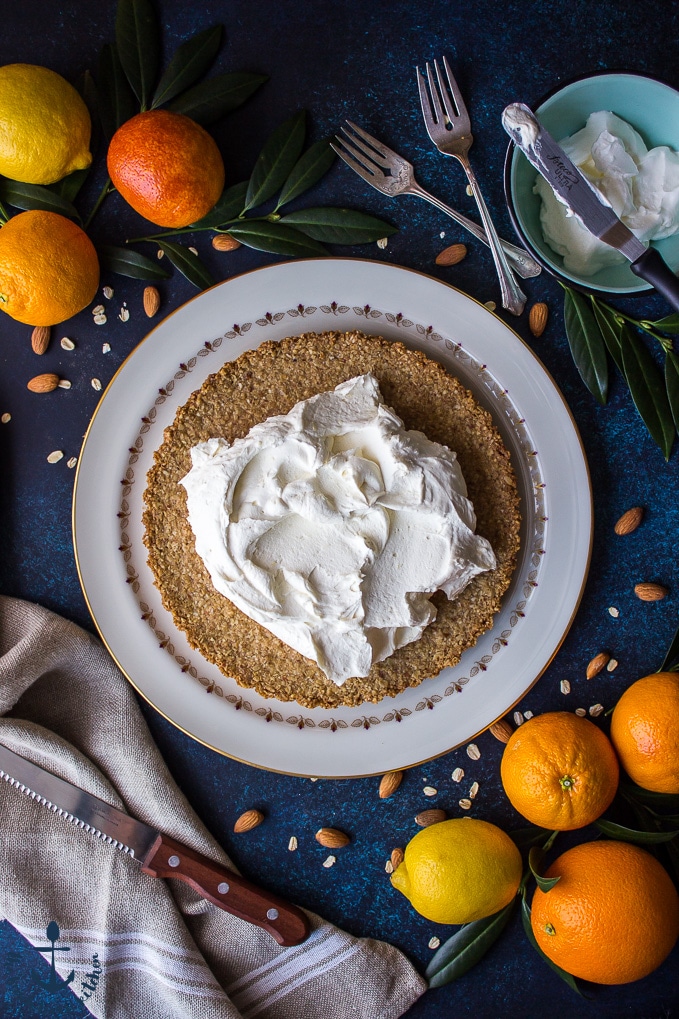 Overhead shot of tart crust topped with mascarpone cream on round white plate with oranges, lemon, sharp knife, forks and bowl filled with whipped cream in background