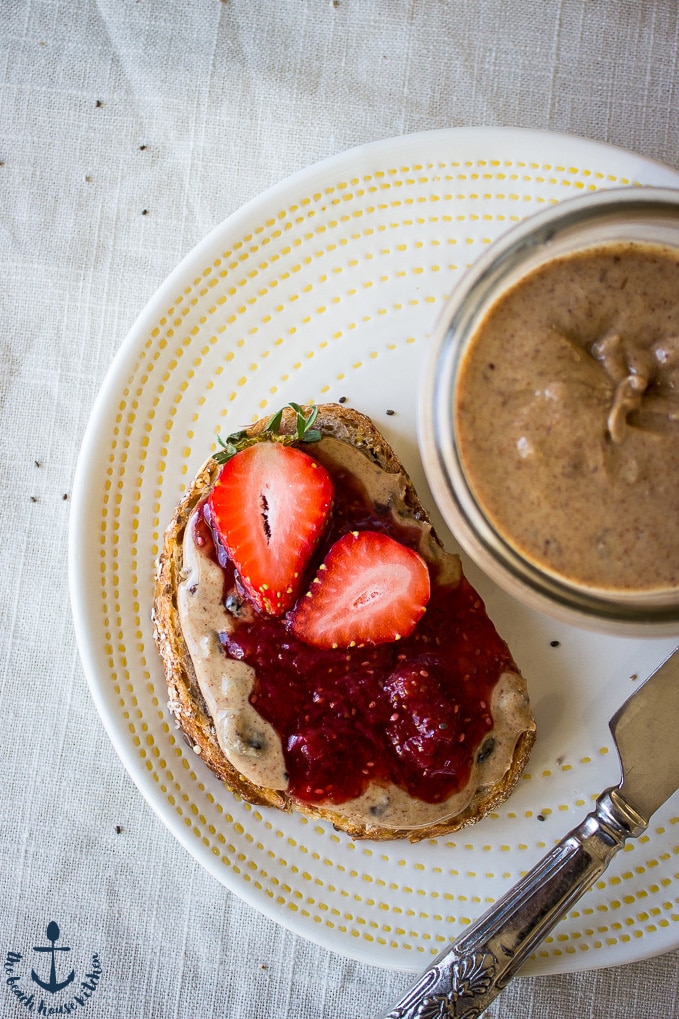 An overhead photo of an open face piece of bread with Cinnamon Raisin Almond Butter and Strawberry Chia Jam with an open jar of almond butter on a white plate with a silver knife.