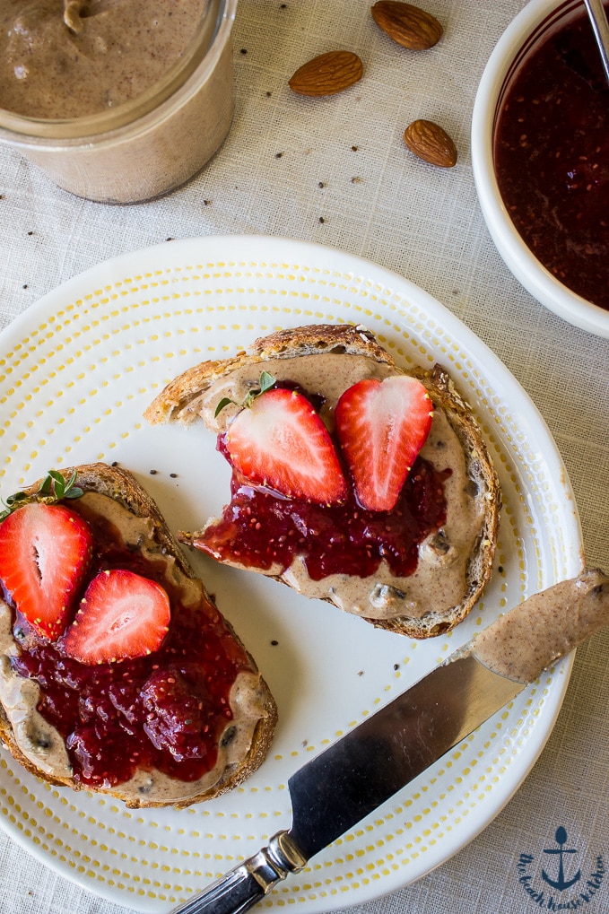 An overhead photo of an open face sandwich of Cinnamon Raisin Almond Butter and Strawberry Chia Jam with an open jar of almond butter on a white plate with a silver knife.