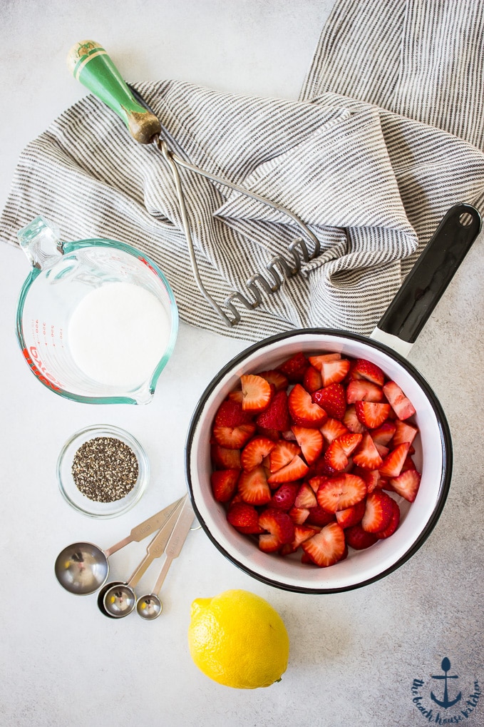 Overhead photo of strawberries in a saucepan, a lemon a potato masher, measuring spoons and measuring cup with a striped dish cloth.