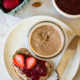 An overhead photo of an open face piece of bread with Cinnamon Raisin Almond Butter and Strawberry Chia Jam with an open jar of almond butter, a container of strawberries and a bowl of strawberry jam.