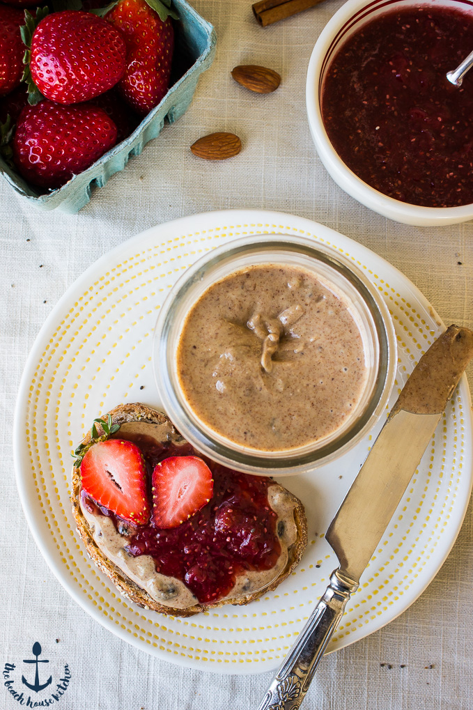 An overhead photo of an open face piece of bread with Cinnamon Raisin Almond Butter and Strawberry Chia Jam with an open jar of almond butter, a container of strawberries and a bowl of strawberry jam.