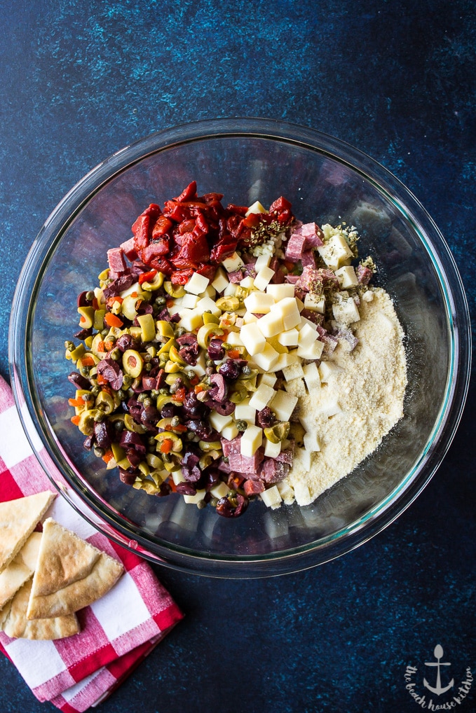 Overhead photo of Muffuletta Dip ingredients in a glass bowl on a blue background with a red and white checked napkin.