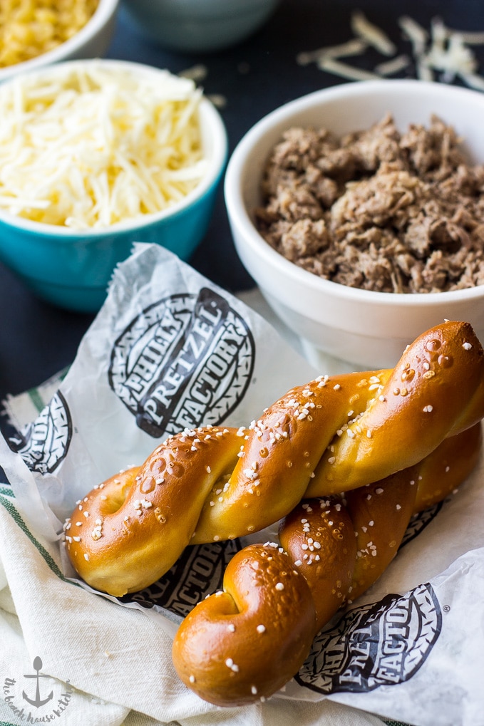 Two Philly soft pretzels with a bowl of shredded cheese and a bowl of cooked steak meat.