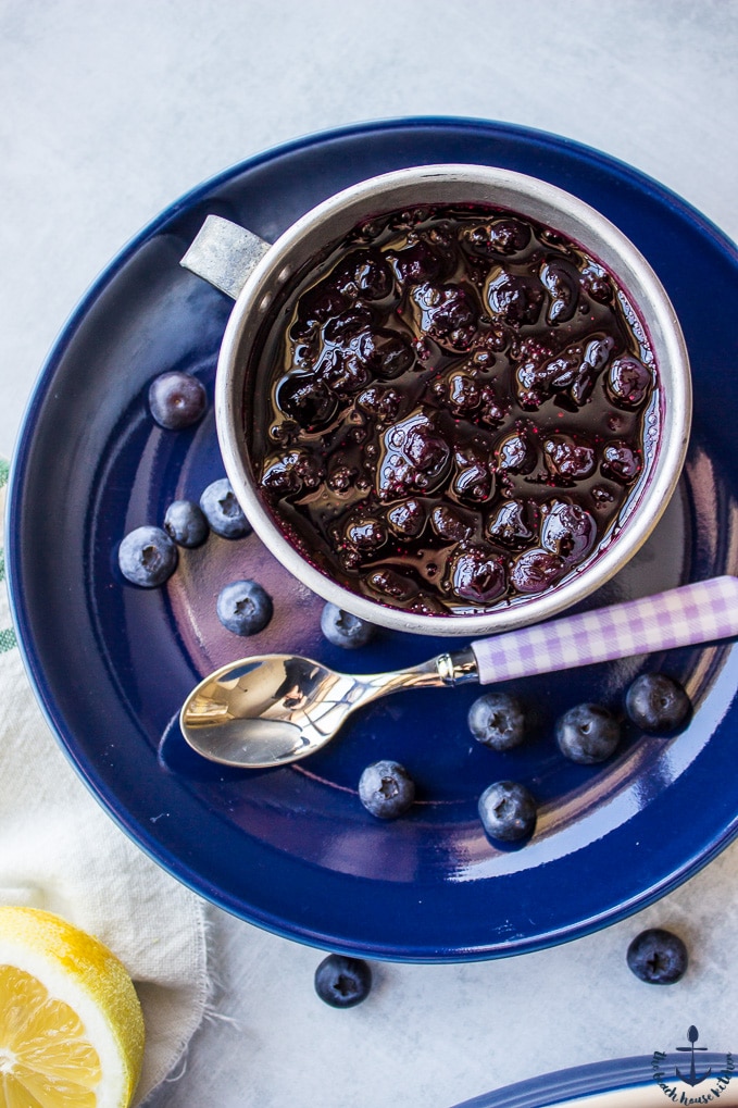 Overhead photo of blueberry sauce in a silver cup on a blue plate with a spoon.