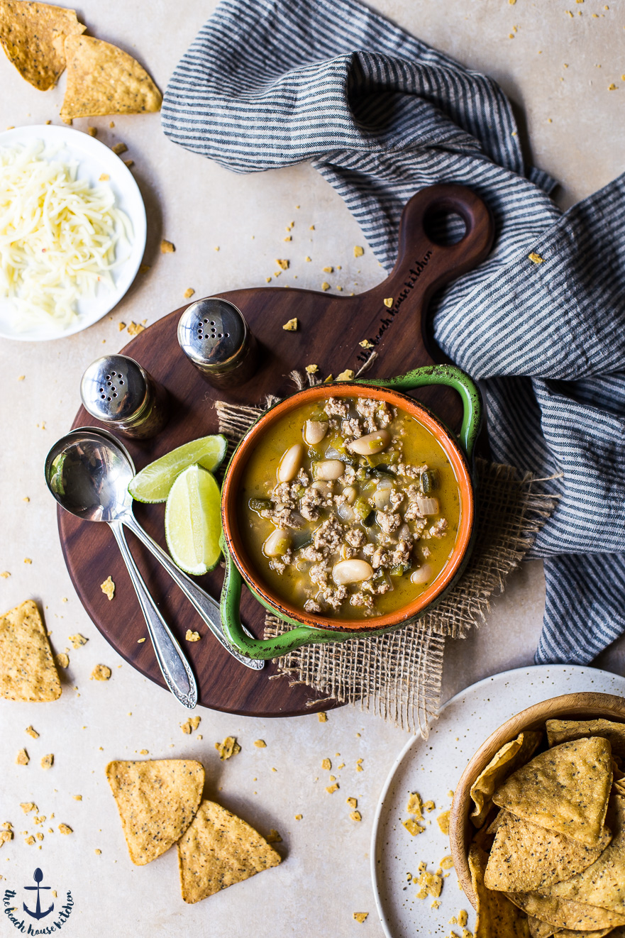 A small bowl of White Bean Turkey Chili on a wooden board with spoons, limes and tortilla chips