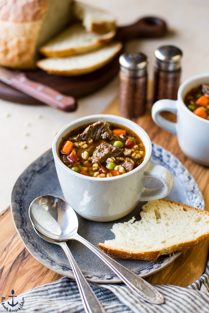Beef Barley Soup in a mug on a blue plate with two spoon and a slice of bread