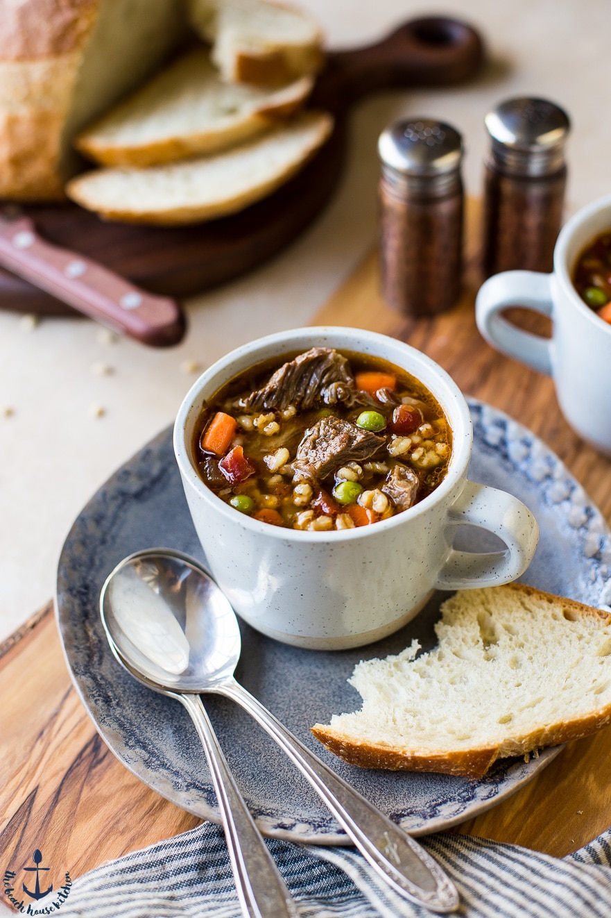 Up close photo of a cup of beef barley soup on a plate with spoons and bread