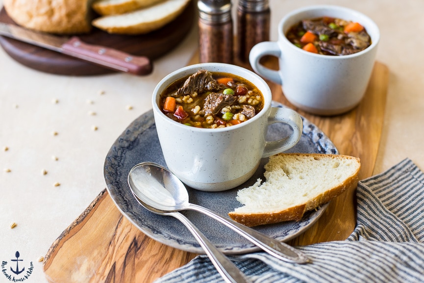 Beef Barley Soup in mugs with one on a plate with spoons and bread