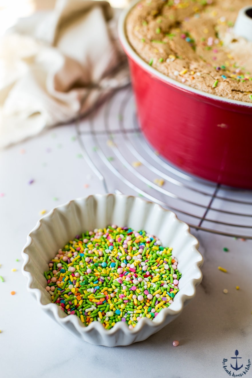 A white bowl filled with sprinkles with an angel food cake in background