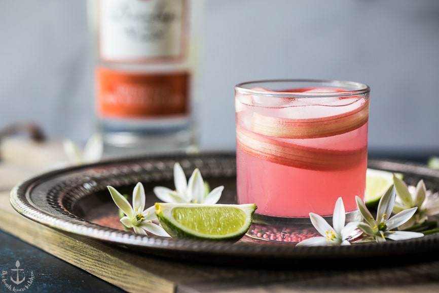 Photo of a pink spiced rhubarb collins cocktail on a silver tray surrounded by flowers and limes.