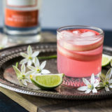 Photo of a pink spiced rhubarb collins cocktail on a silver tray surrounded by flowers and limes.